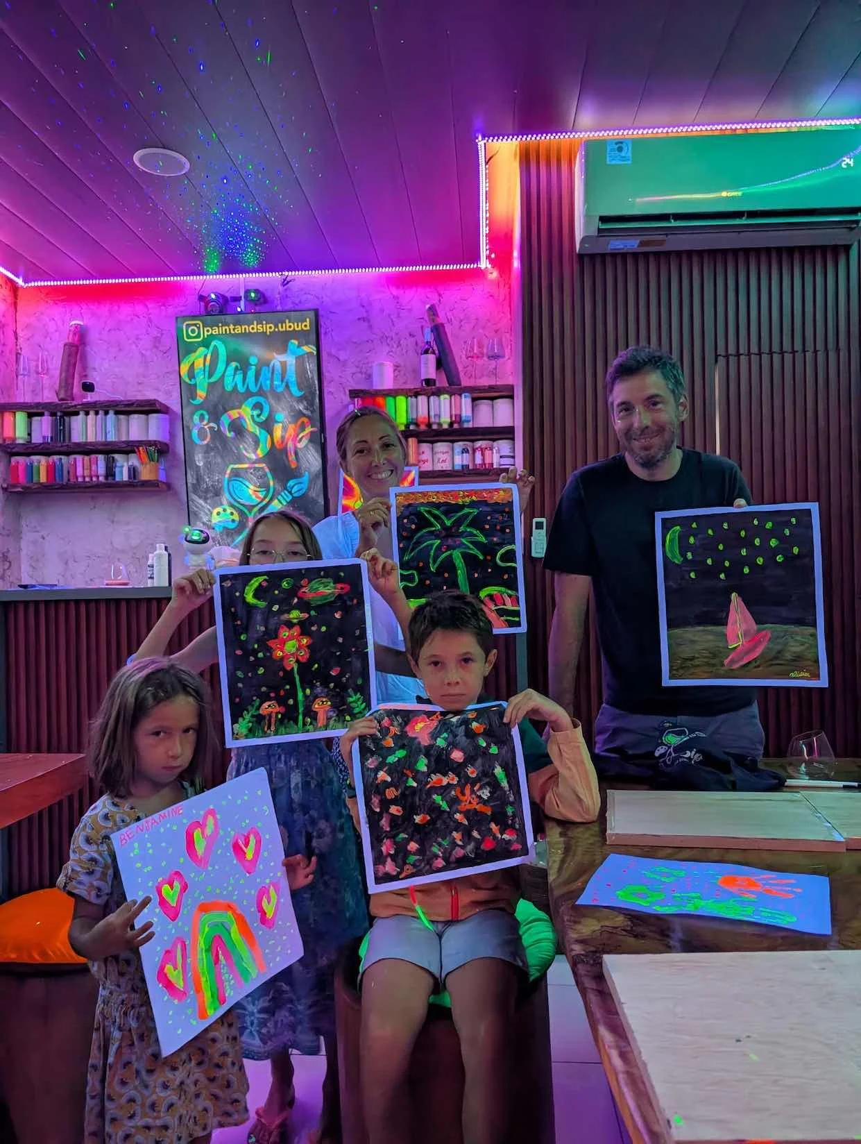 Group of children and two adults in a bright, colorful art studio holding up neon painted artwork, with a 'Paint & Sip' sign on the wall, surrounded by neon lights and art supplies.