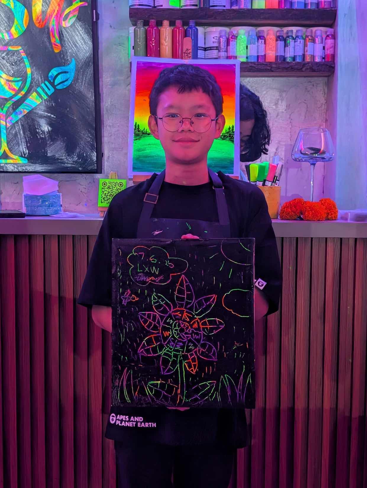 A young man with glasses wearing a black apron, holding a light-up drawing of a flower and leaves on a black background, in a colorful art studio with neon and rainbow lighting.