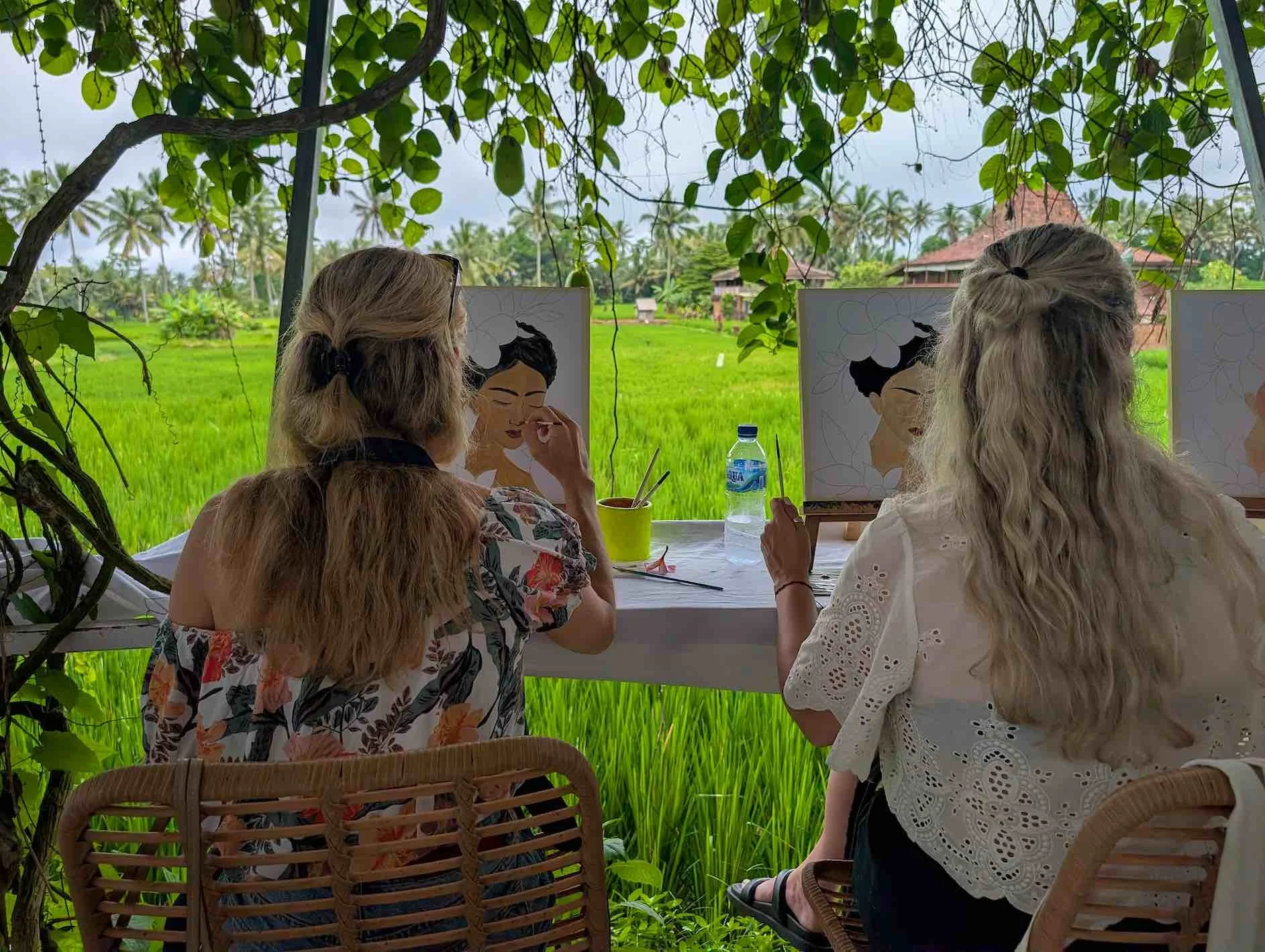 Two women painting portraits of women with dark hair and glasses, sitting at a table outdoors with green fields and palm trees in the background.