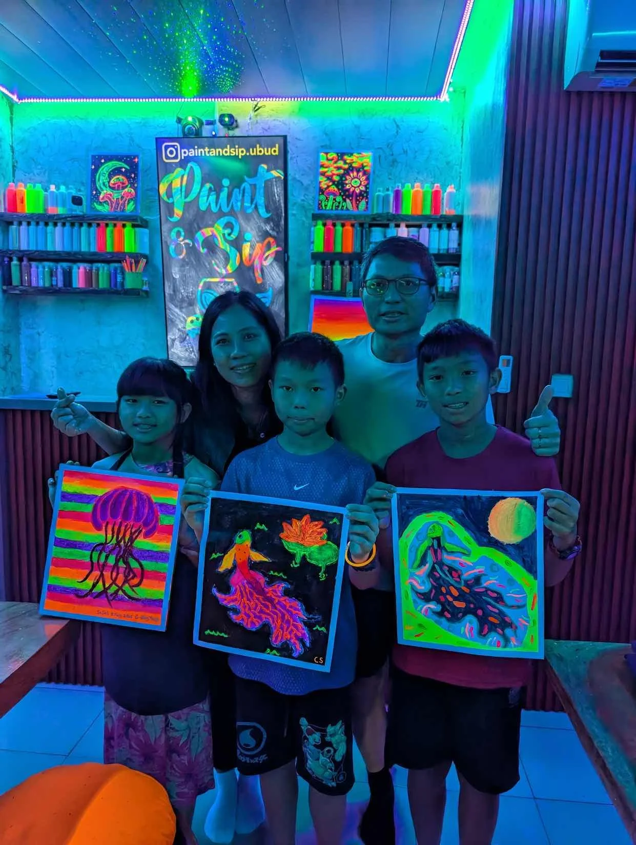 Group of five people holding neon glow-in-the-dark paintings in an paint and sip studio with black lights and colorful wall art.