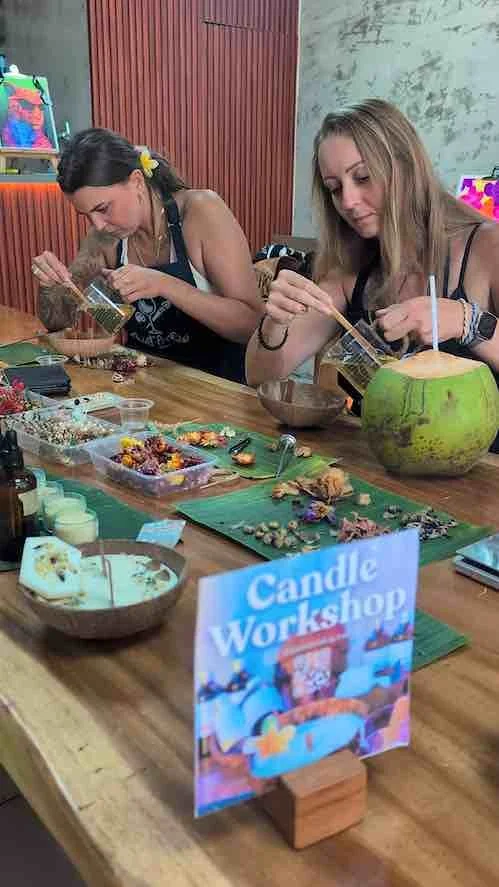 Two women creating candles at a woodworking workshop, with various craft supplies on the table and a sign reading 'Candle Workshop' in the foreground.