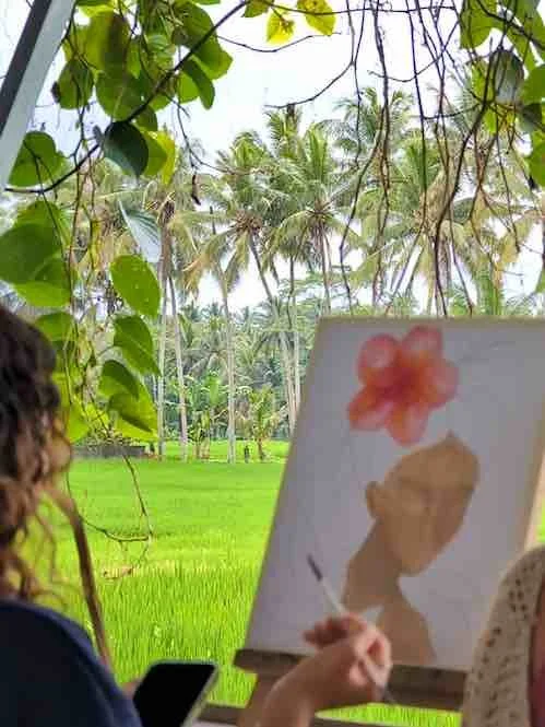 Person painting a portrait of a woman with a red flower in her hair outdoors, surrounded by lush greenery and palm trees.