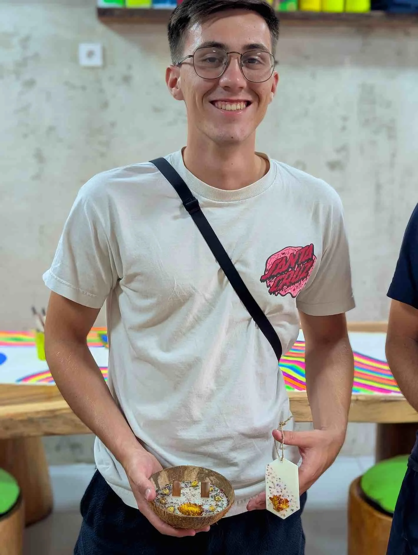 A smiling young man with glasses wearing a white T-shirt with a pink graphic, holding a wooden bowl filled with small flowers and a tag, standing indoors in front of a wall with colorful artwork.
