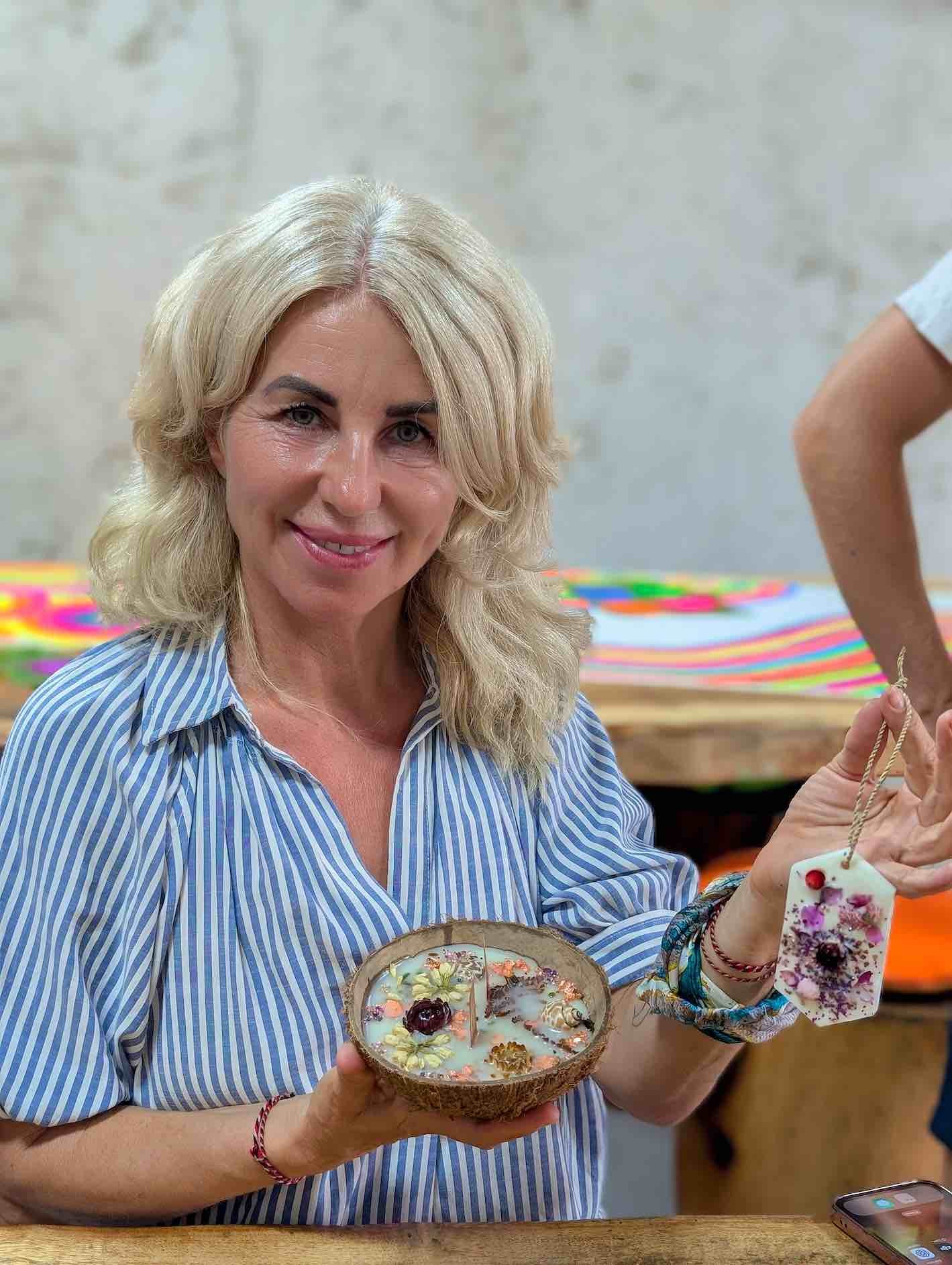 Woman with blonde hair holding a decorated candle and a handcrafted soap, sitting at a table with colorful objects in the background.