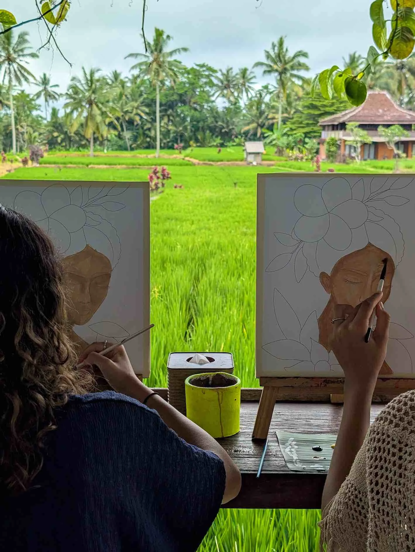 Two women are painting portraits of women on canvases, with a lush green landscape and palm trees visible through the window behind them.