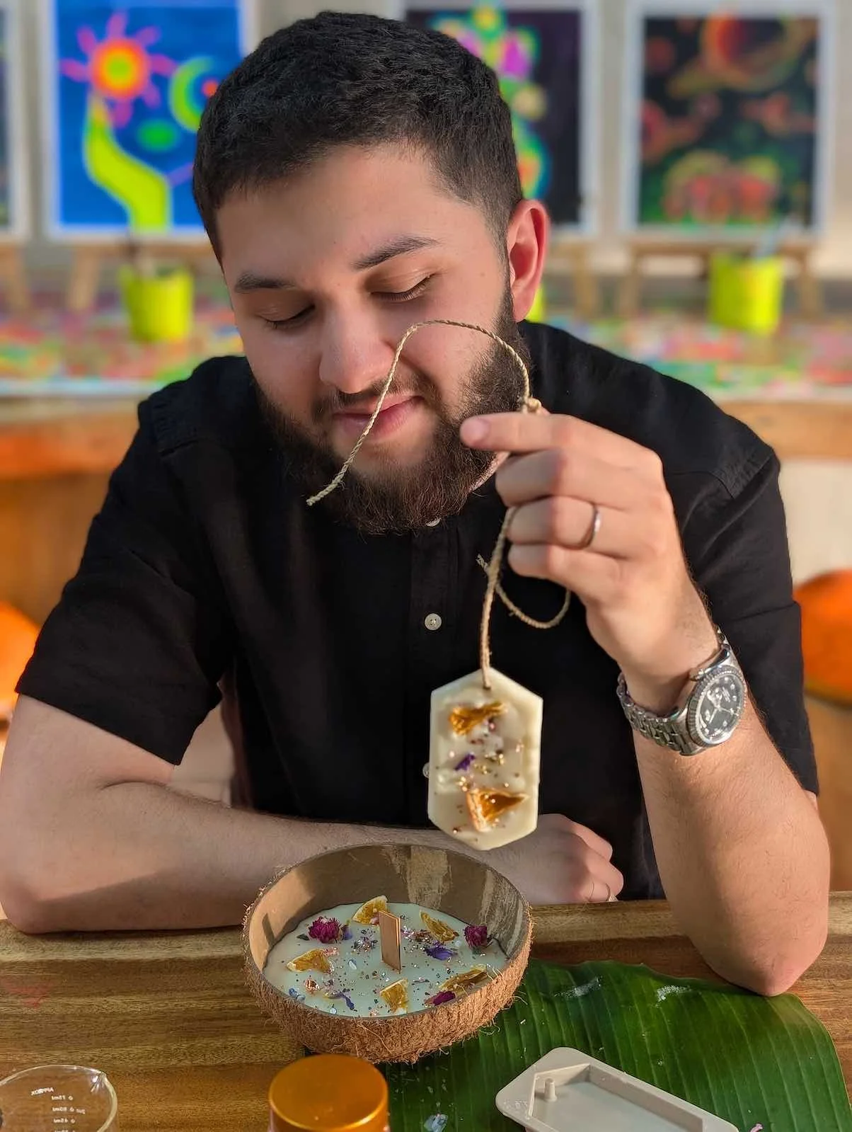 A man with a beard, wearing a black shirt, holds a handmade soap or decorative ornament hung on a string, with colorful dried flowers embedded, over a bowl containing similar flowers on a table decorated with a large green leaf. Background features c