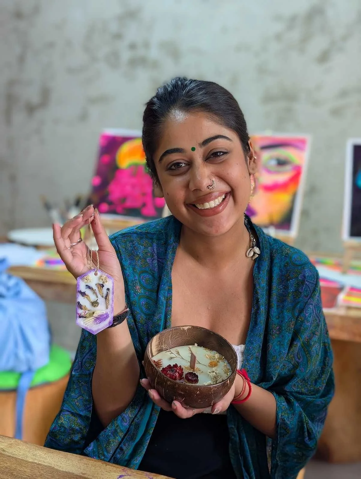 A woman smiling and holding a brown bowl with a candle and berries, wearing a blue shawl and jewelry, standing in an art studio.