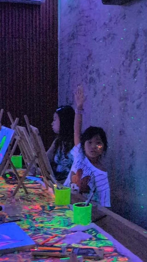 Two children, a girl and a boy, participating in a glow-in-the-dark painting activity at a table covered with neon-colored paint and art supplies, with a wall in the background illuminated by blacklight.