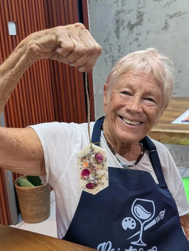 An elderly woman with short white hair, smiling, holding a decorative piece of soap with dried flowers and herbs, wearing a dark apron over a white shirt.