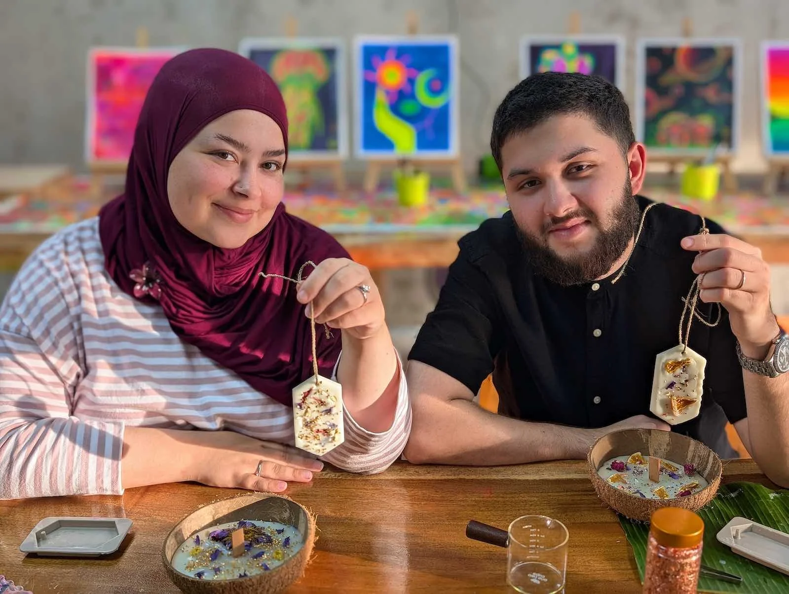 Woman and man smiling and holding candle tags in a craft workshop with art on the wall behind them.