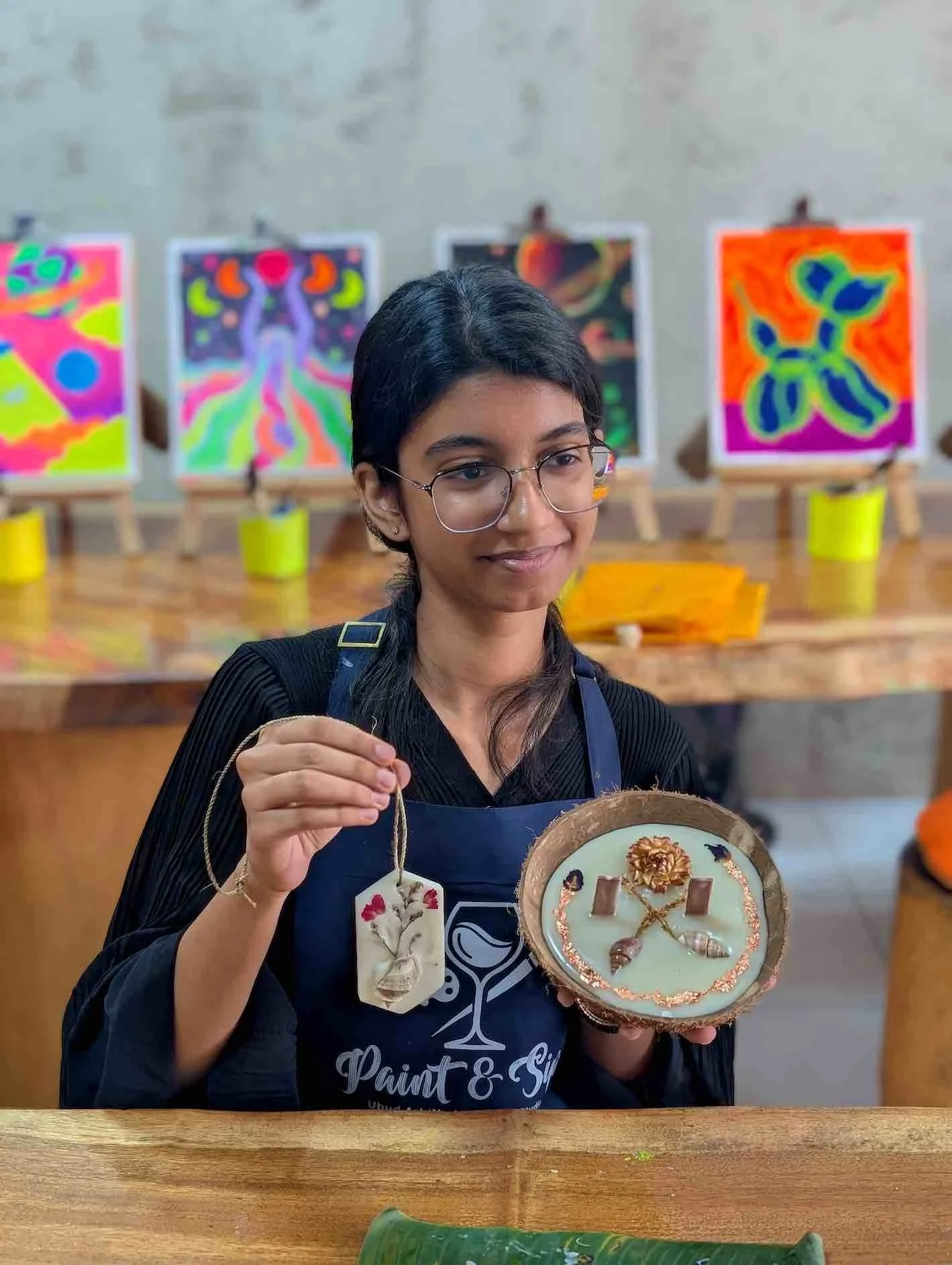 A woman holding a decorated art piece made of wood and ceramic, in a studio with colorful abstract paintings in the background.