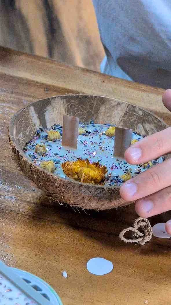 A handmade candle inside a coconut shell bowl, decorated with dried flowers and small candles, with two wooden sticks standing upright. The bowl is on a wooden surface.