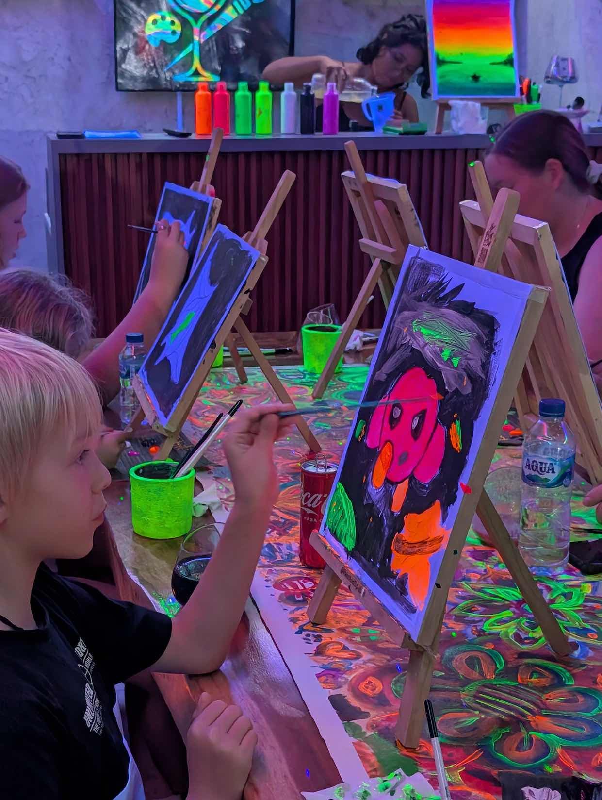 Children and adults painting colorful pictures on small canvases at a glow-in-the-dark painting party, with neon paints and black lights creating a luminous effect.
