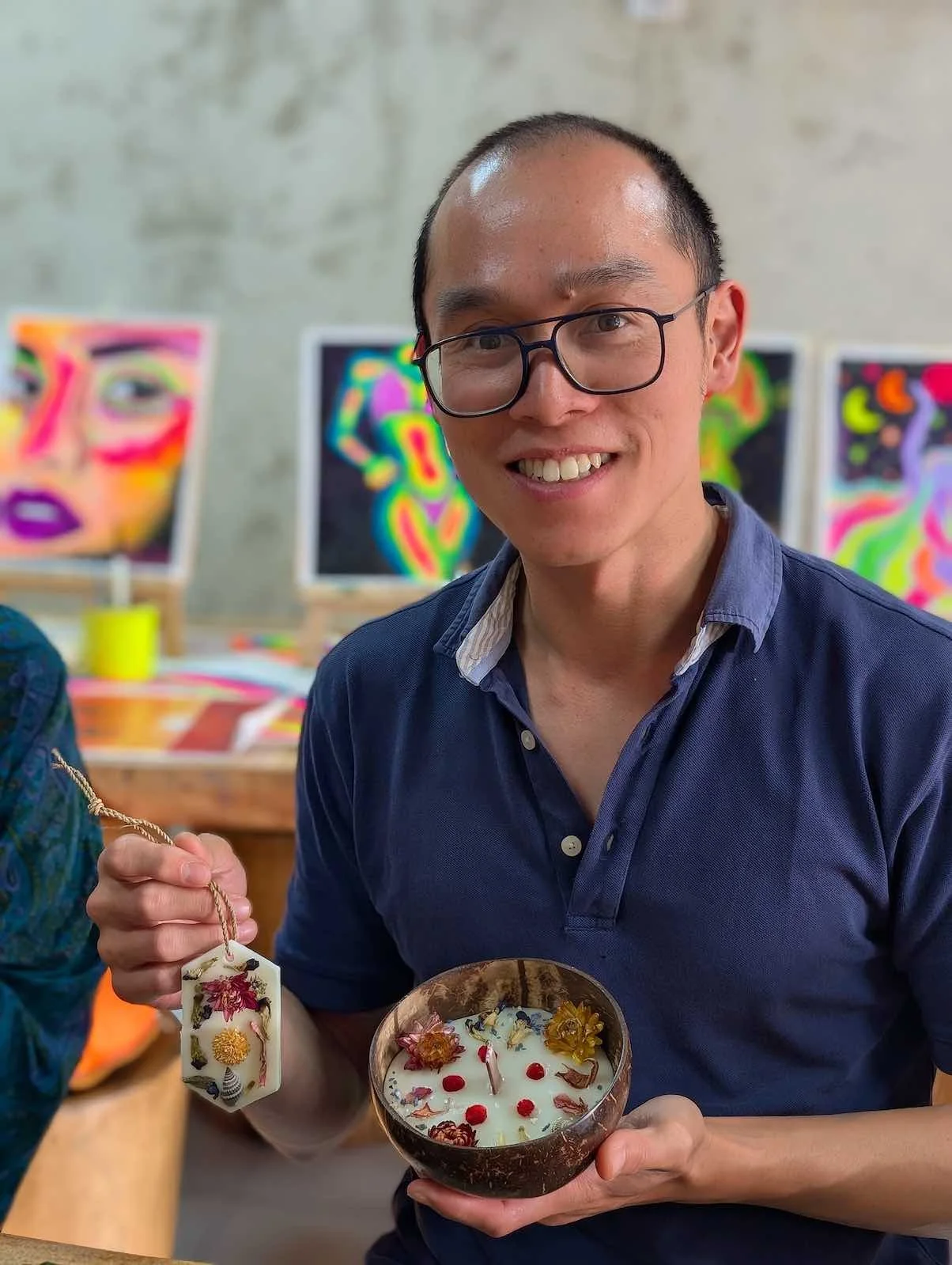 A smiling man with glasses, wearing a blue shirt, holding a decorative bowl with a candle inside and a tag with floral designs, in an art gallery with colorful abstract artwork in the background.