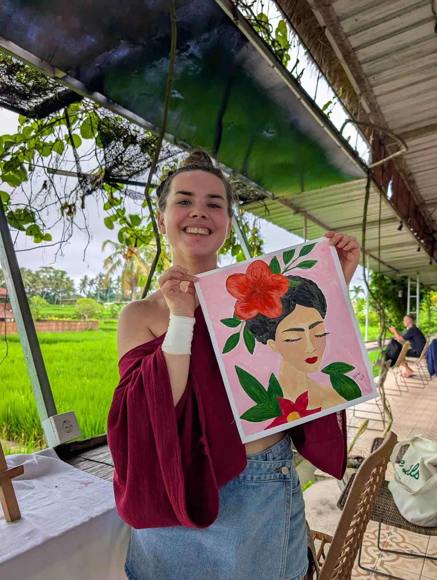 A young woman with a smiling expression holding a painting of a woman with an updo hairstyle, red flowers, and green leaves. She is wearing a burgundy off-the-shoulder top and a denim skirt, standing outdoors with lush green fields and a few people i