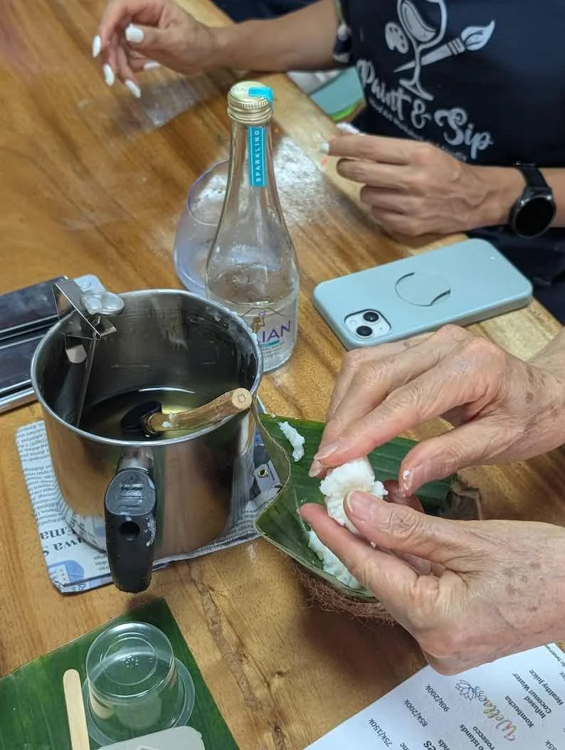 A person preparing coconut with a spoon, a pot of boiling water, a glass bottle, and a smartphone on a wooden table.