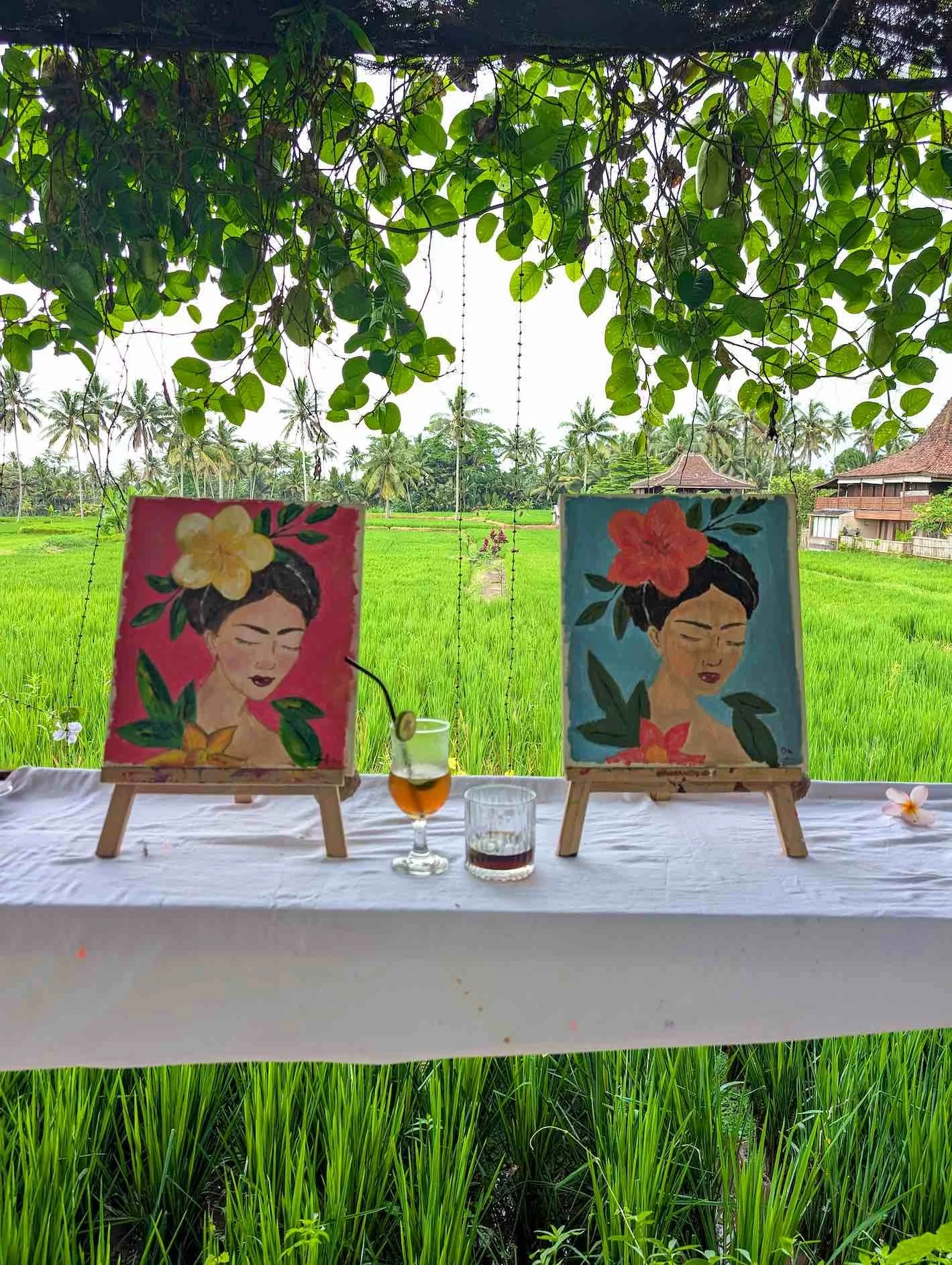Two paintings of women with flowers in their hair on small easels, placed on a table with drinks, set against a lush green rice field under a canopy of leaves.