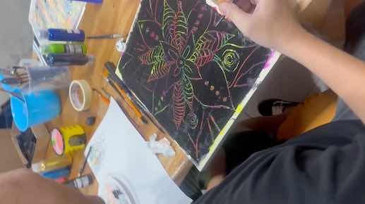 A person working on a colorful chalk drawing of a large butterfly on a black surface, with various art supplies like chalk pastels, tape, and paper on a wooden table.