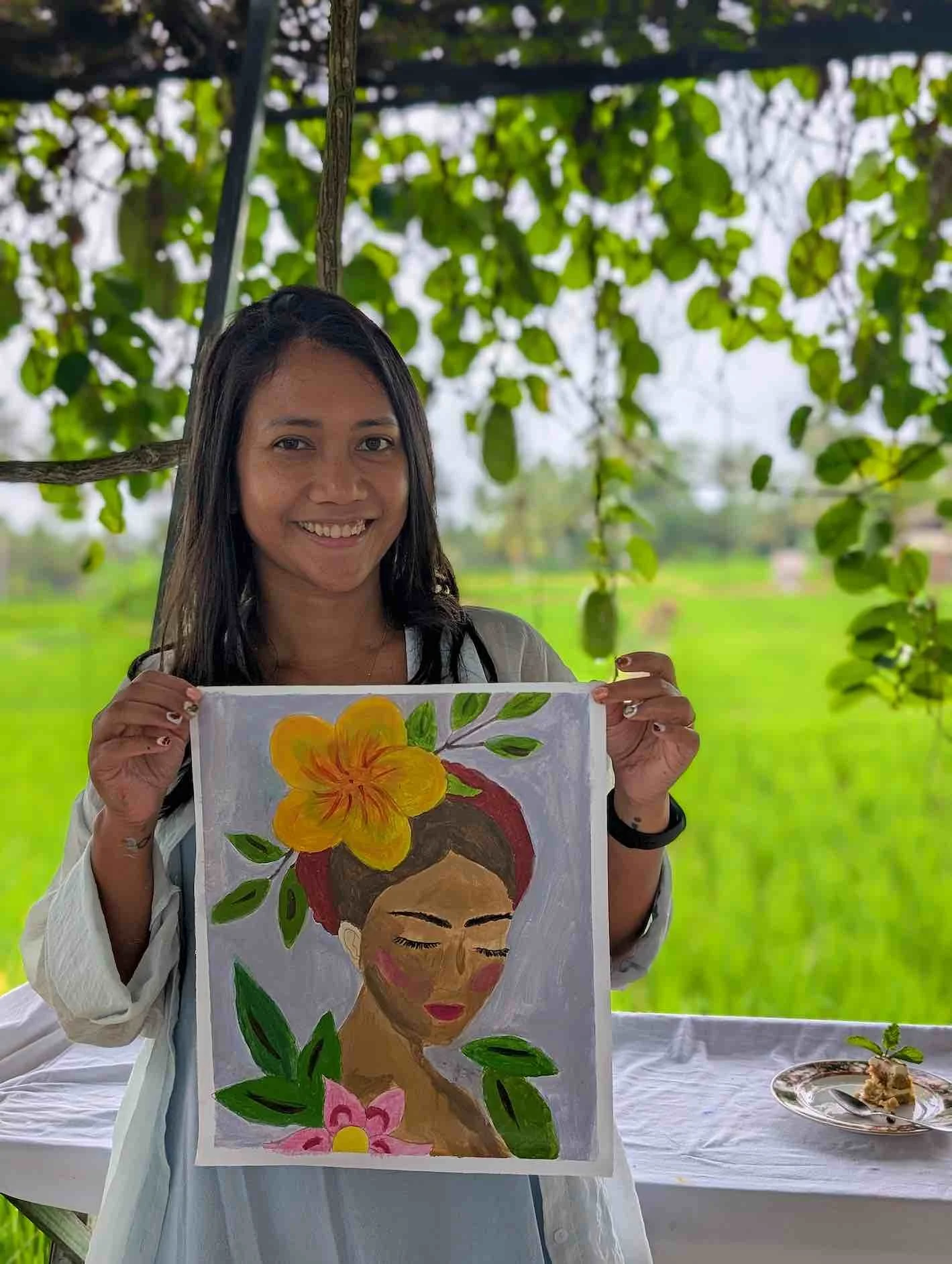 A woman smiling outdoors holding a colorful painting of a woman with flowers and leaves, with a green field in the background.