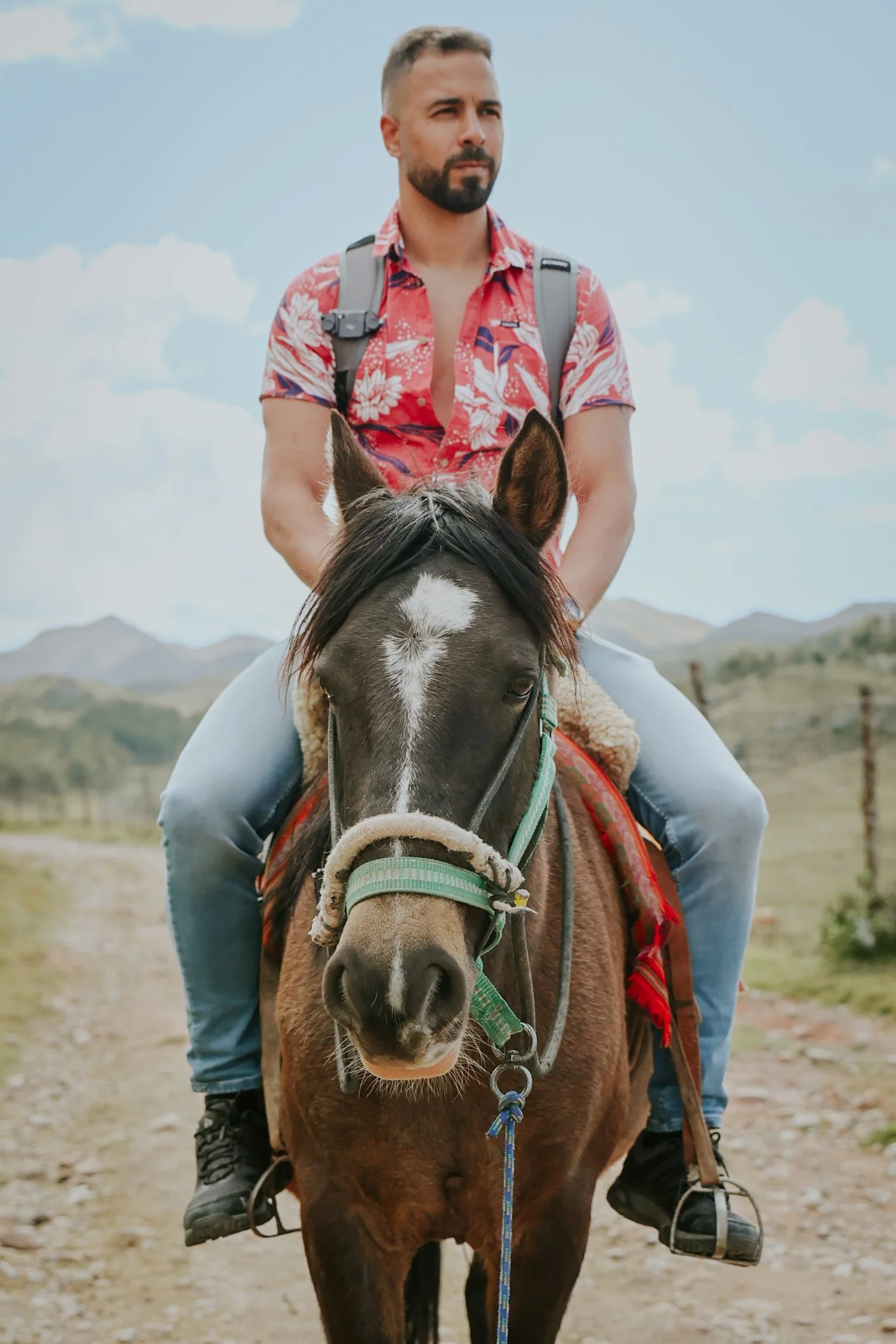 Un homme avec un sac à dos, portant une chemise hawaïenne rouge et un pantalon en jeans, monte à cheval dans un paysage rural avec des montagnes en arrière-plan.