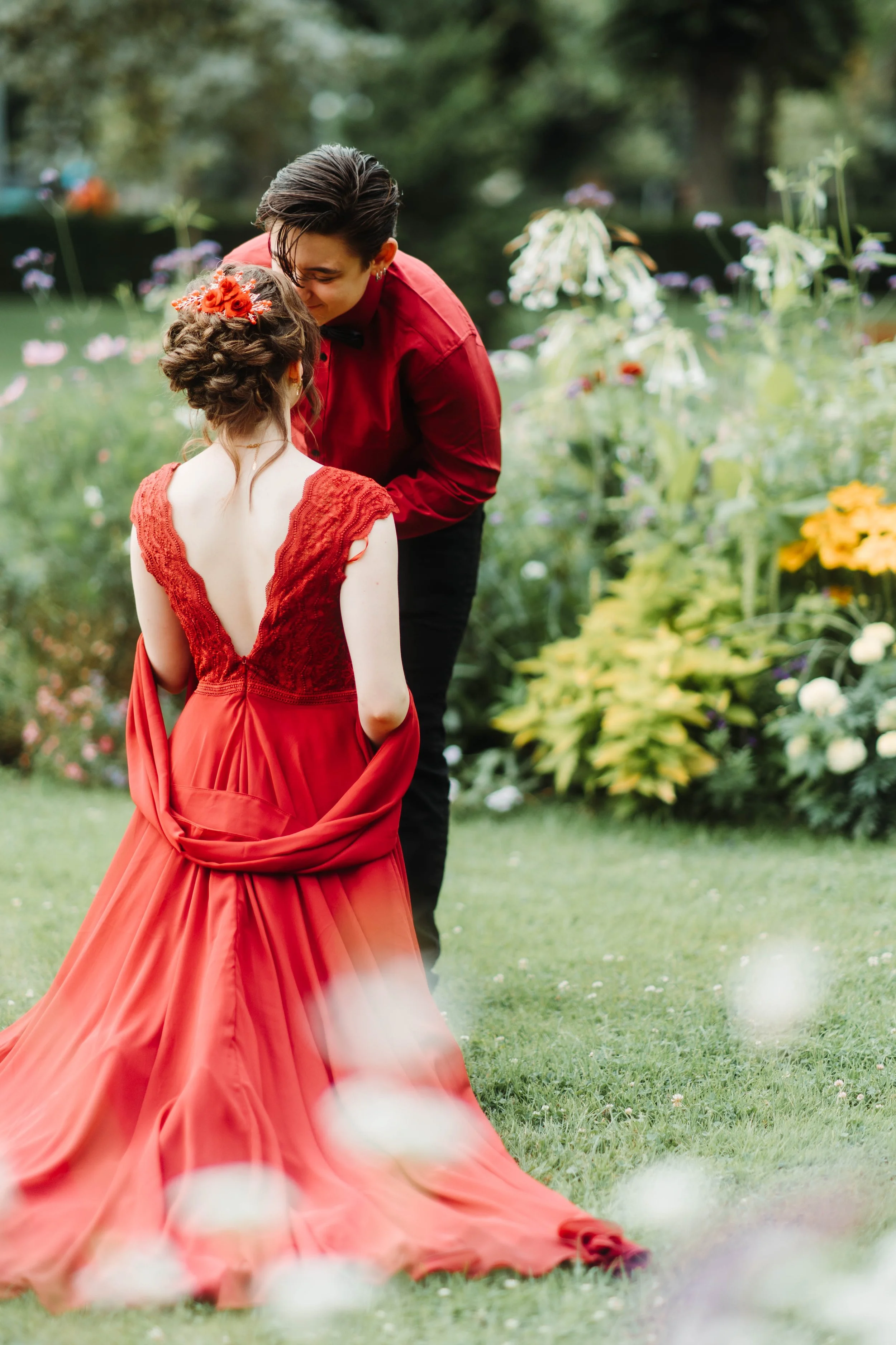 Une femme en robe rouge se penche vers une jeune fille portant une robe rouge avec des fleurs dans ses cheveux, dans un jardin verdoyant.
