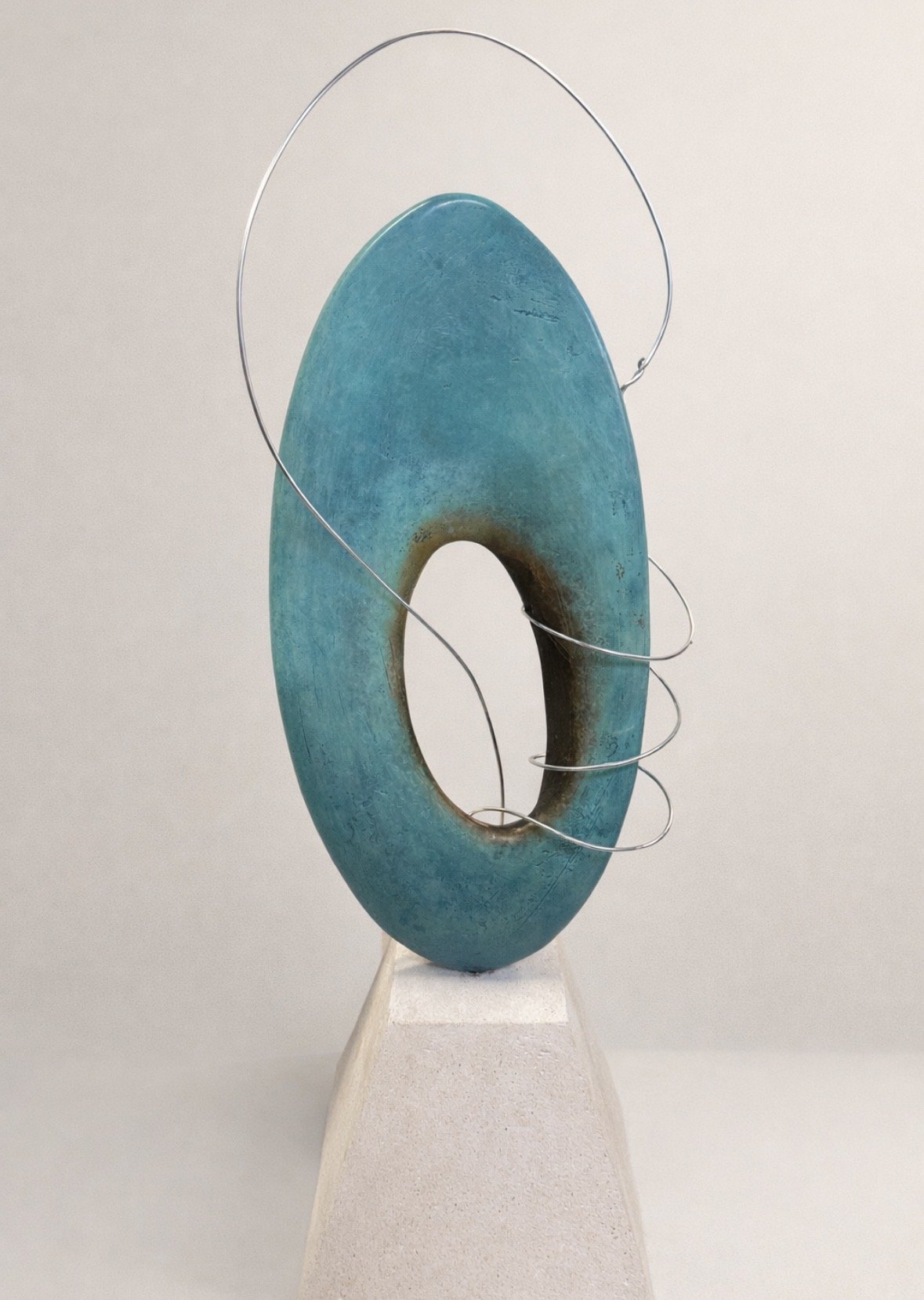 Sculpture of a large turquoise ring with a hollow center, mounted on a white pedestal, with thin metal wire loops around it.