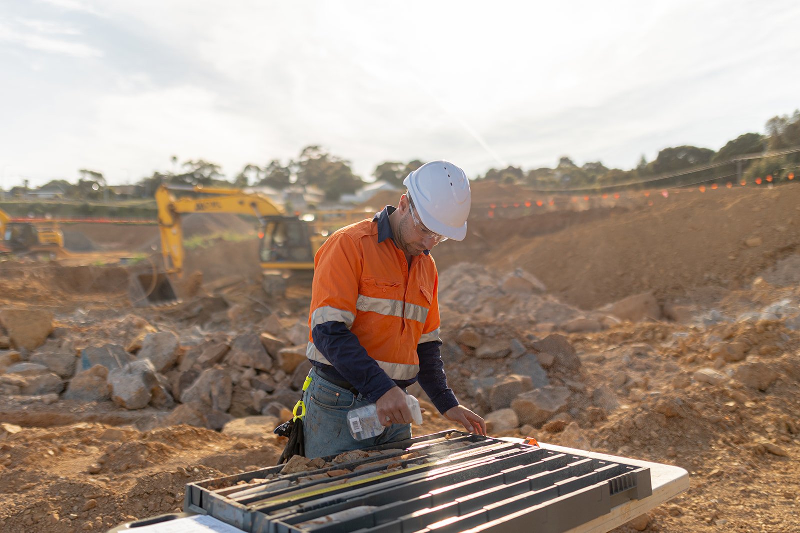 A construction worker in an orange safety jacket and white helmet examines plans at a construction site with excavators in the background.