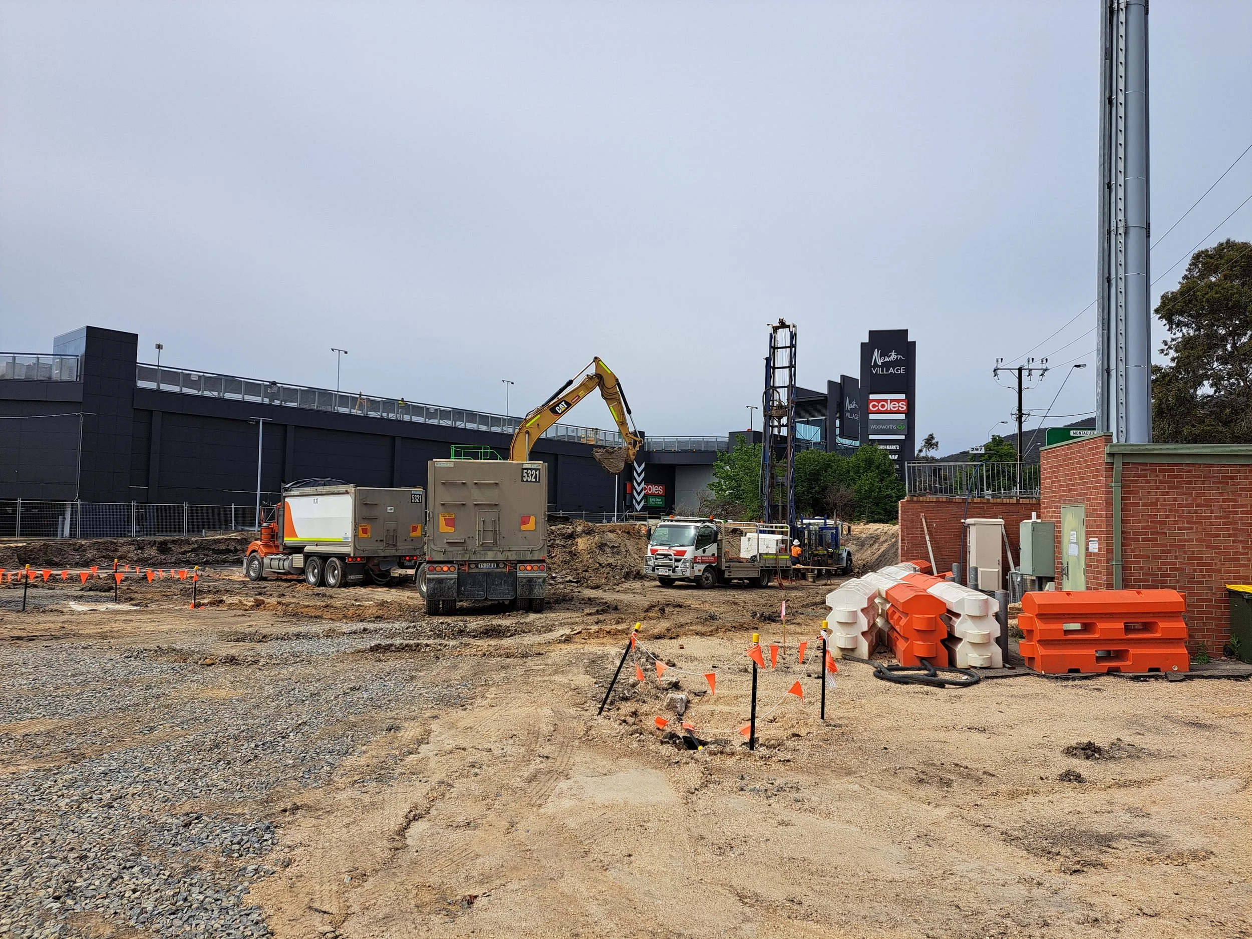 Construction site with excavation equipment and trucks, orange safety barriers, and a brick building wall under cloudy sky, with signage for a shopping center in the background.