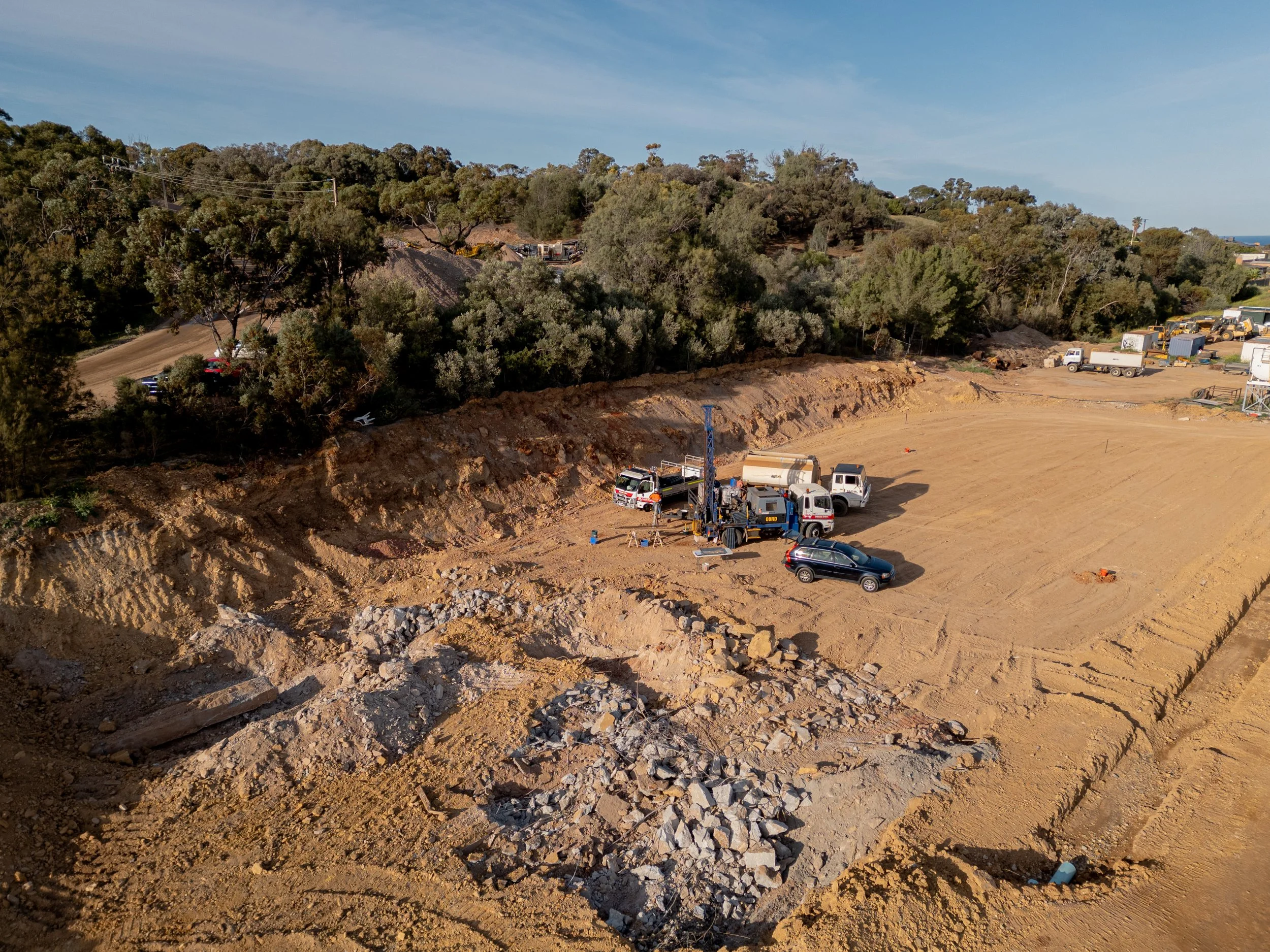 Construction site with drilling equipment, trucks, and excavated ground, surrounded by trees and a clear sky.