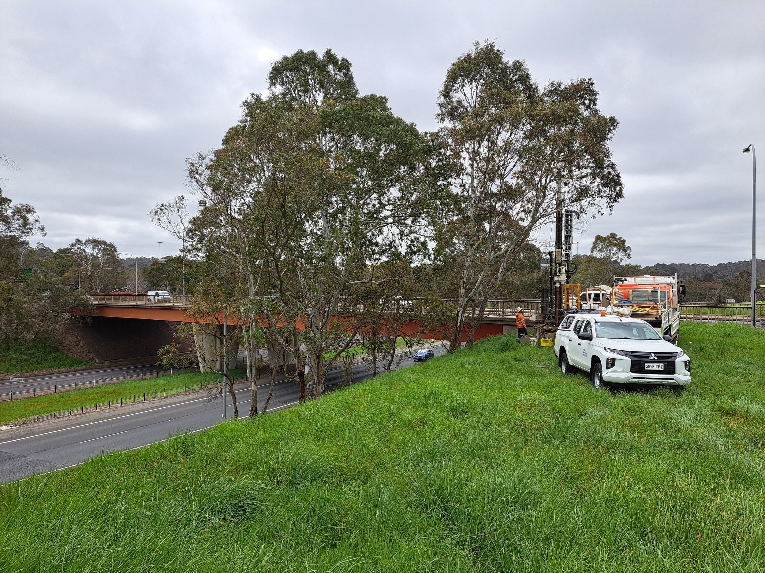 Roadside scene with maintenance trucks and workers fixing traffic signals near a bridge, green grass and tall trees in the background.