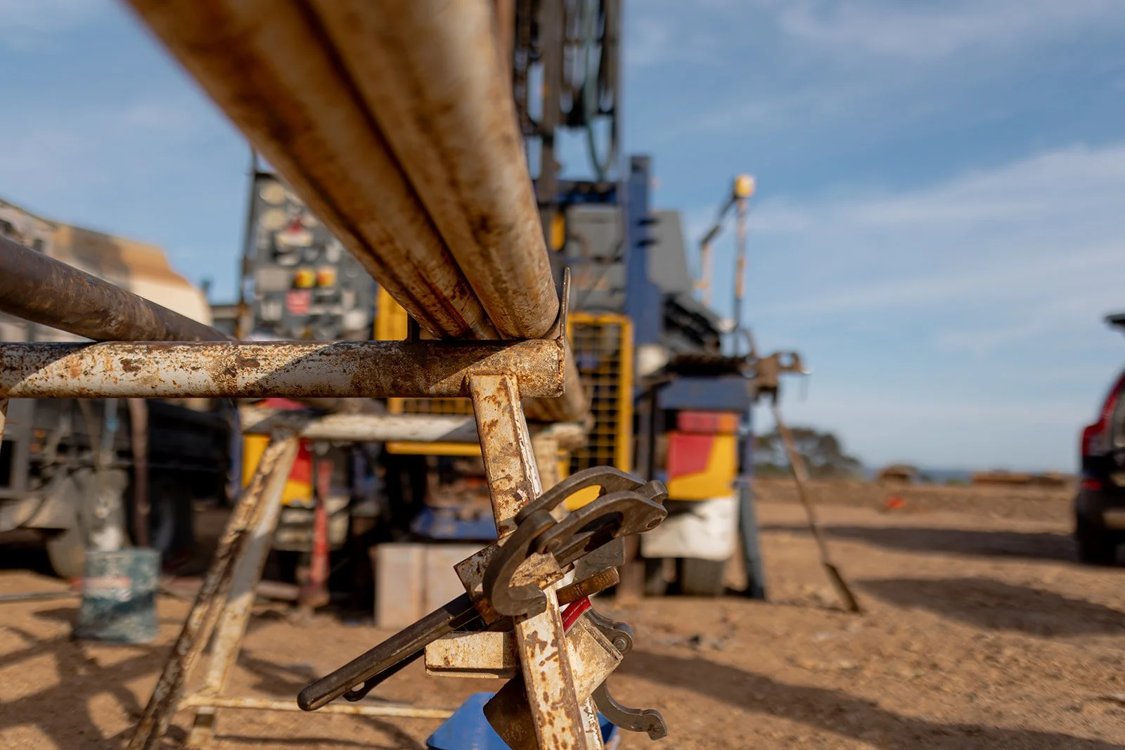 Close-up view of rusted metal pipes and clamping tools in a construction or industrial site with a blurred background of equipment and a partly cloudy sky.