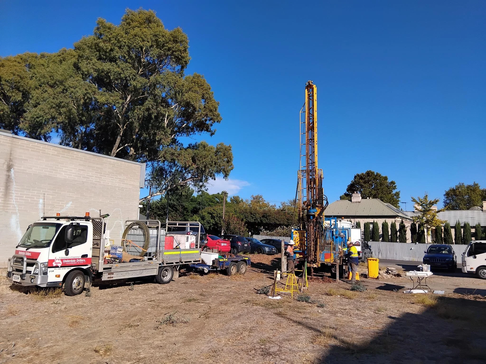 Construction site with workers operating a tall yellow drilling rig, a flatbed truck with equipment, parked cars, a gray building, a large tree, and a clear blue sky in the background.
