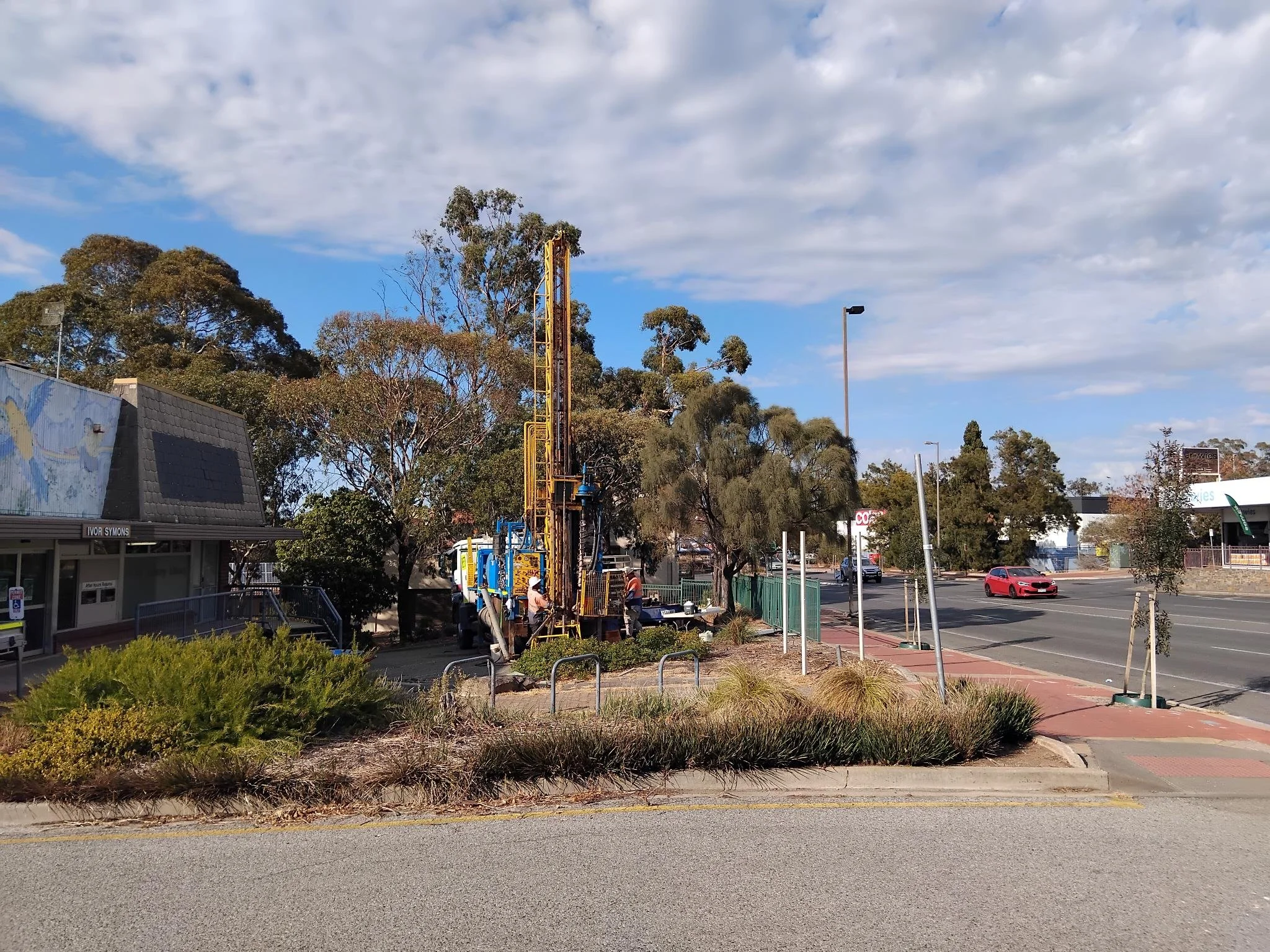 Street view showing a construction vehicle with workers, a sidewalk, trees, and traffic with a red car passing by. Buildings and street signs are visible in the background.