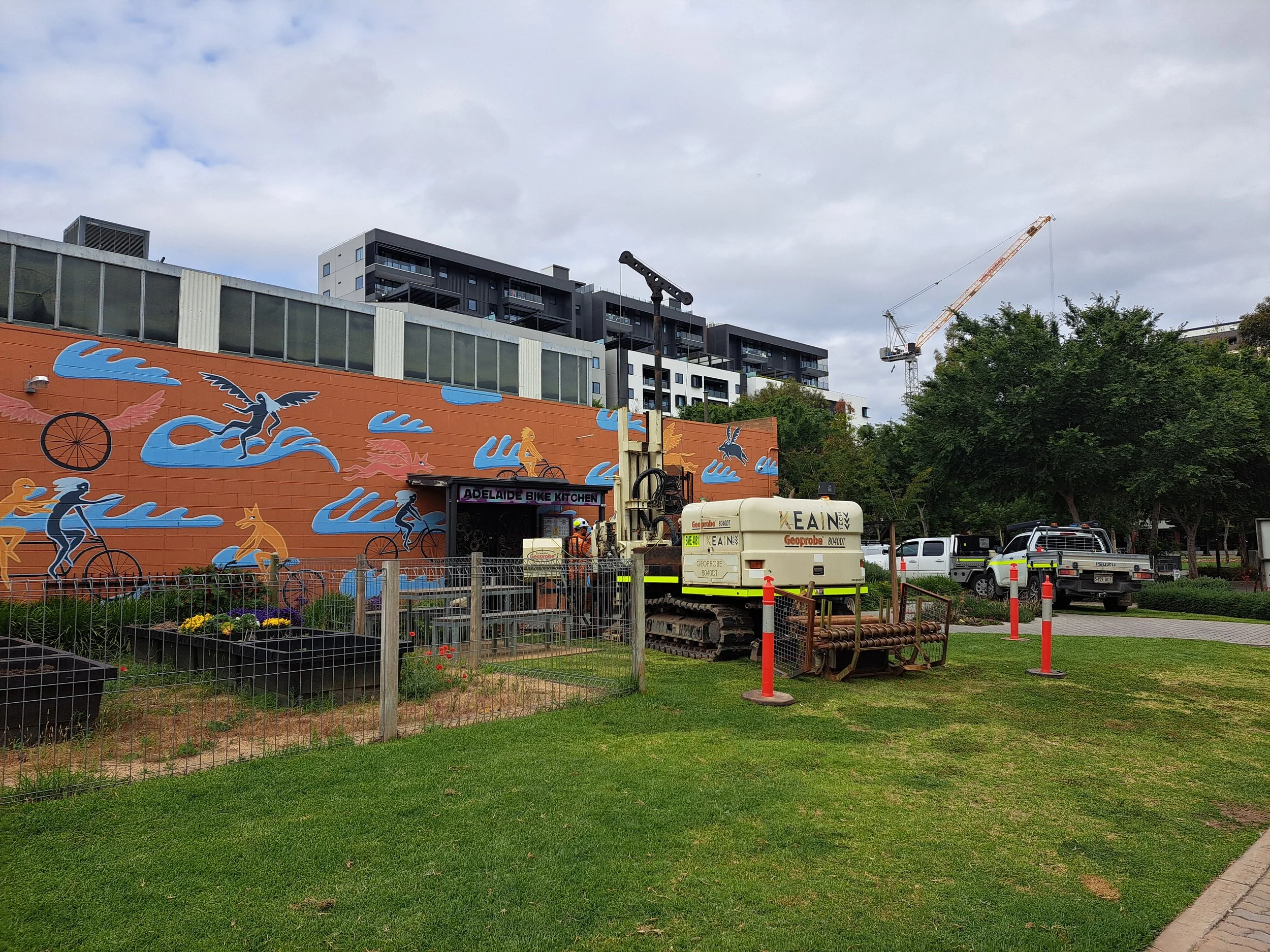 A construction site with a large mural on the wall depicting stylized people and animals riding waves, with modern buildings and a crane in the background. Construction equipment and vehicles are also present.