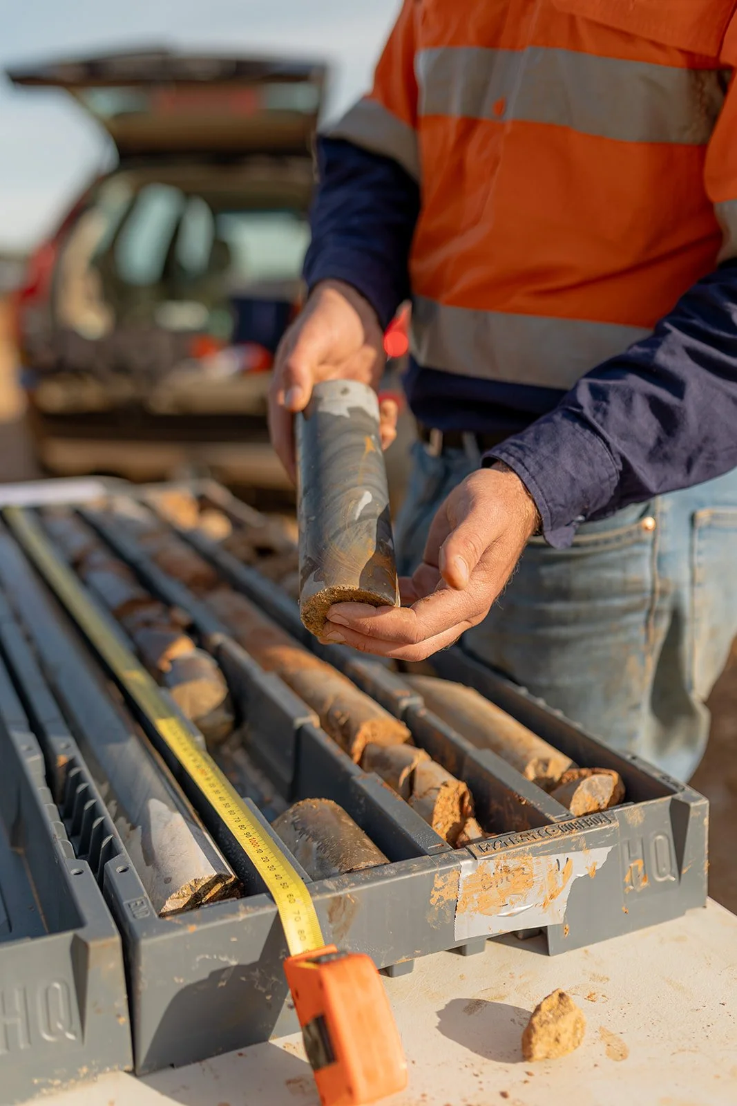 A person in a safety orange and navy blue shirt holding a geological core sample above a toolbox filled with similar samples, with a yellow measuring tape on a work surface.