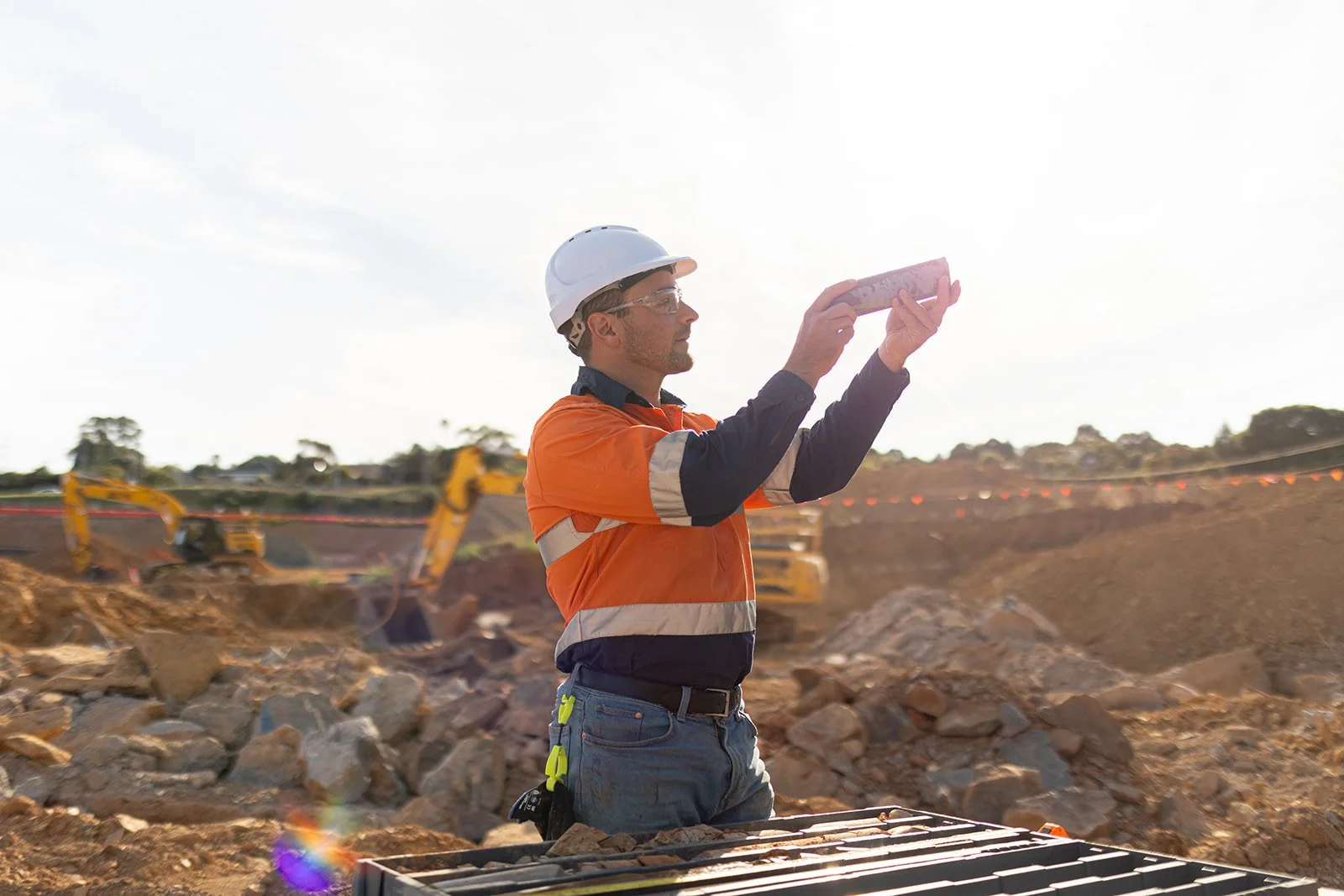 Construction worker wearing a white helmet and orange safety vest taking notes with a clipboard at a construction site with excavators in the background.