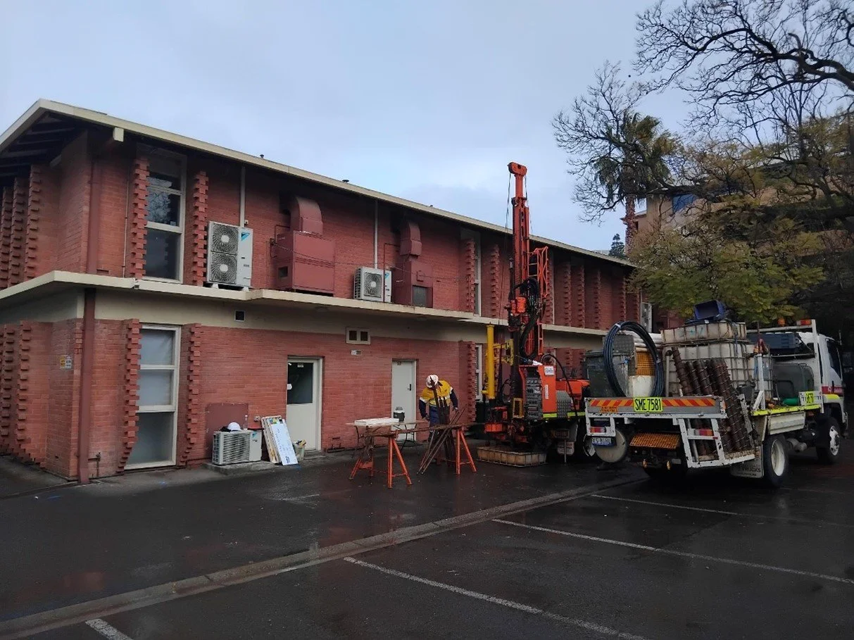Construction worker operating drilling equipment outside a red brick building on a rainy day.