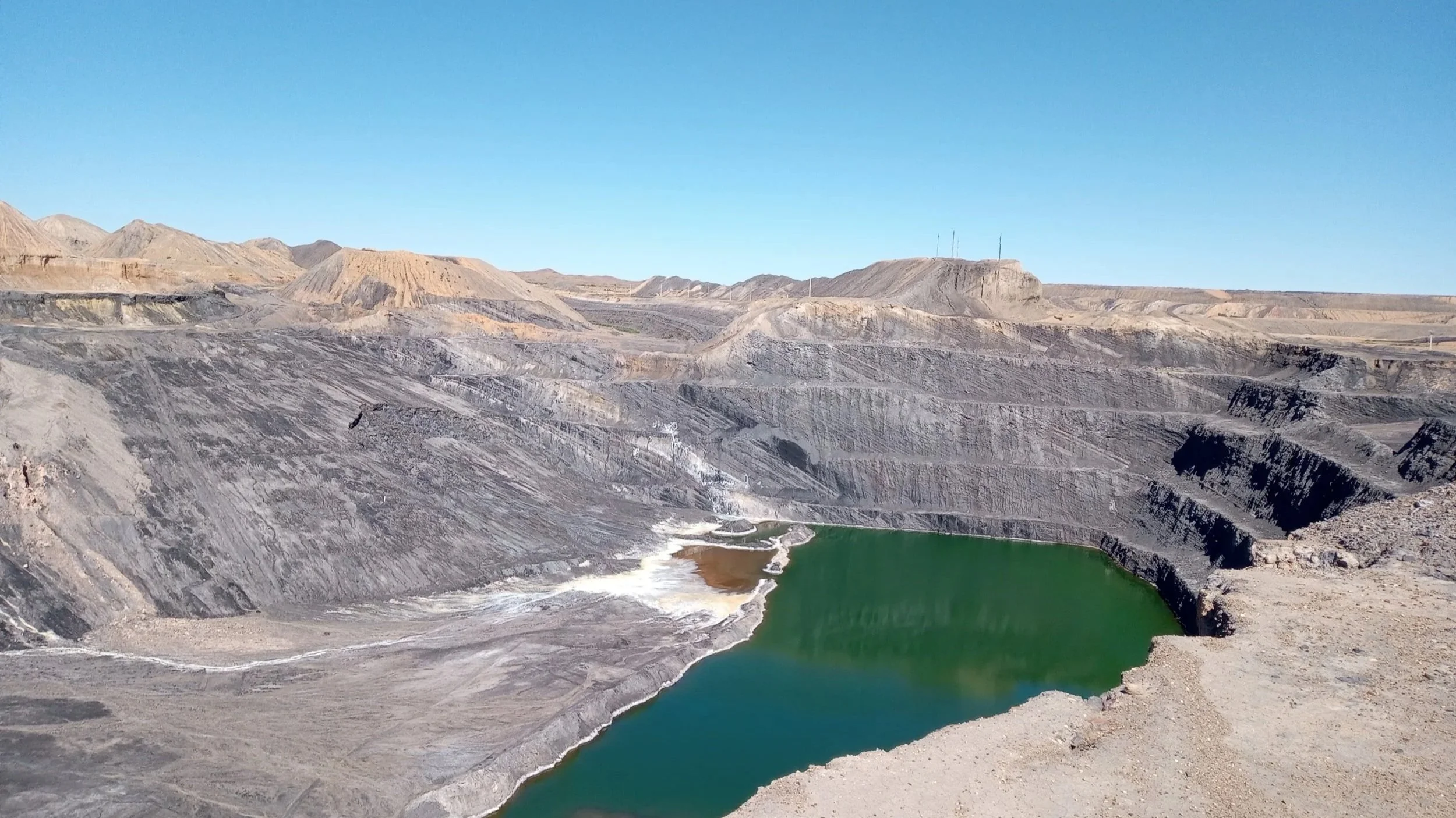 A barren, rocky landscape with a deep quarry and a small body of water at the bottom, under a clear blue sky.