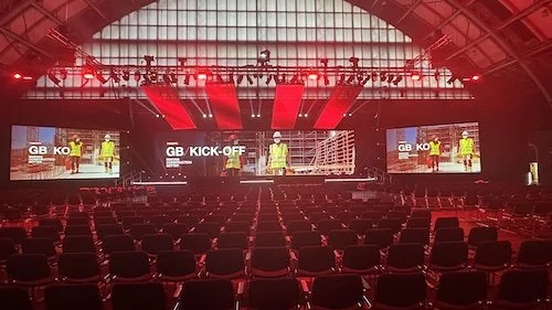 Indoor conference hall with a large stage, two screens displaying construction workers, and red lighting overhead, prepared for an event.
