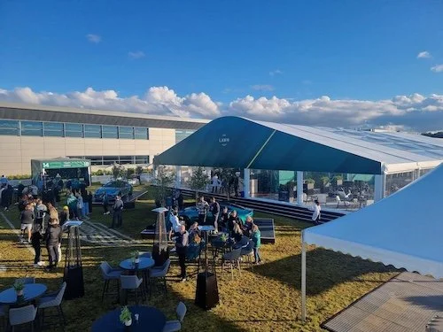 Outdoor event with people gathered near tents and a building labeled 'The Lawn' under a blue sky with clouds.