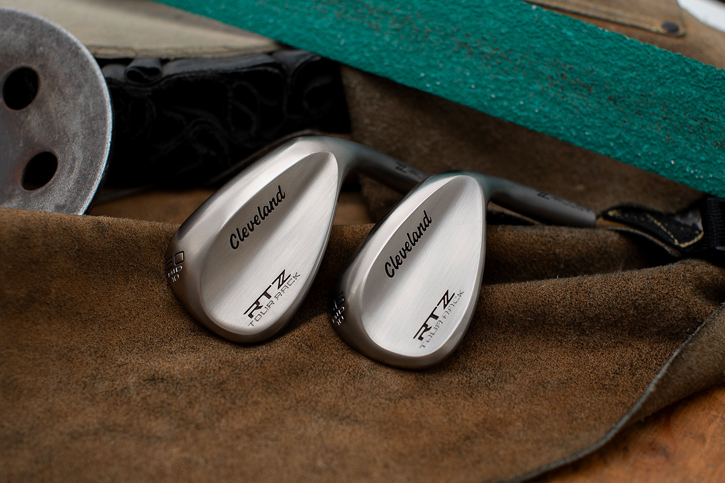 Two Cleveland RTX Tour Rack golf wedges with a brushed metal finish resting on a brown cloth, with a green foam pad, a rusted metal disc, and a black strap in the background.