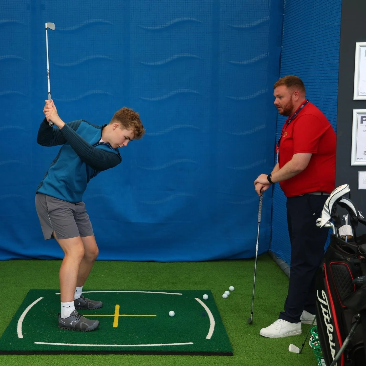 A young man practicing golf swings in an indoor golf simulator with an instructor observing, standing on a green mat with golf balls around.
