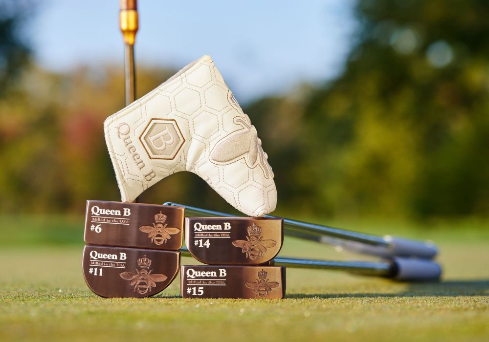 A golf club with a white headcover resting on top of four boxes of Queen B golf balls, on a golf course with a blurred background of trees.