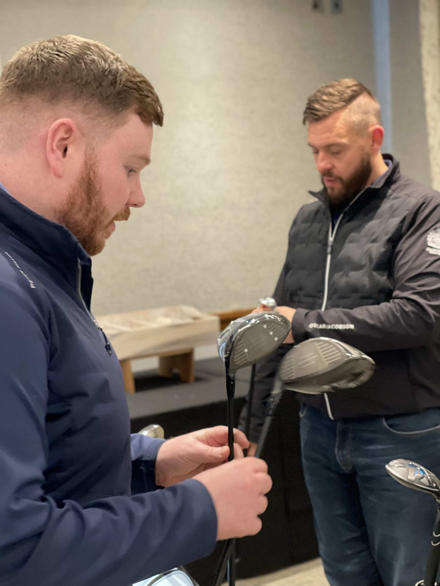 Two men inspecting golf clubs indoors.