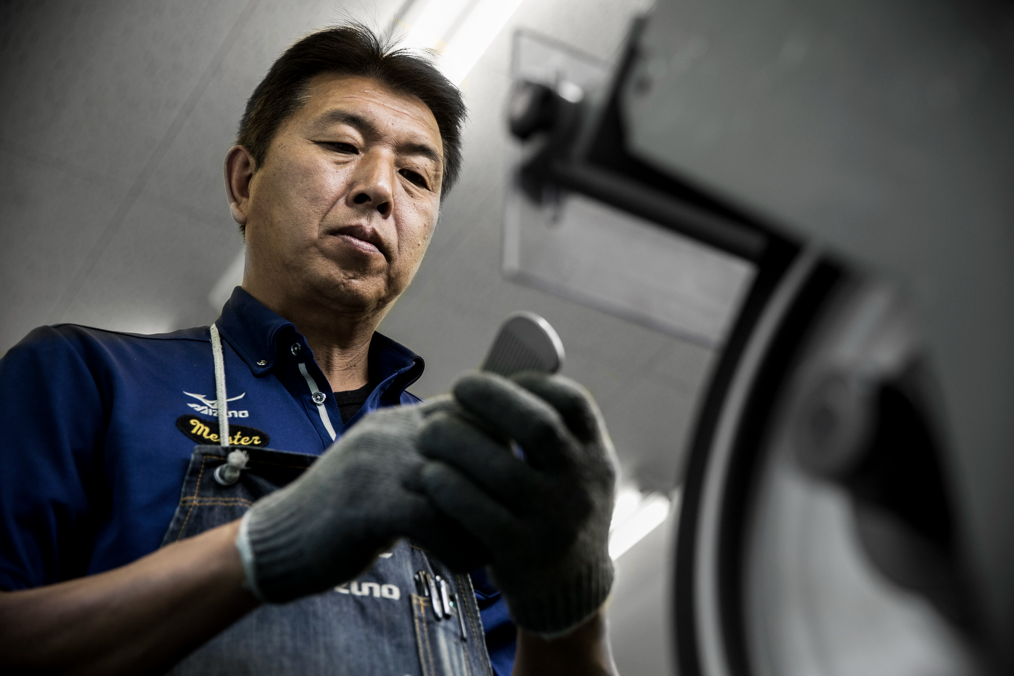 A mechanic wearing a blue polo shirt and gray gloves examining a part of a car.
