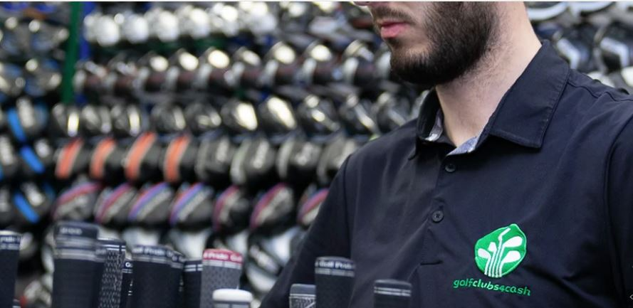 A man in a navy polo shirt looking at golf balls in a sporting goods store, with golf clubs displayed in the background.