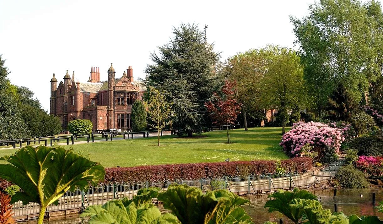 A large park with green grass, variety of trees, and blooming bushes in front of a historic brick building.