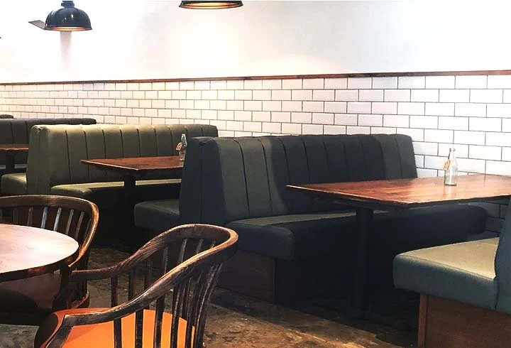 Empty restaurant with dark green and black booths, wooden tables, and black pendant lights, white brick wall in background.
