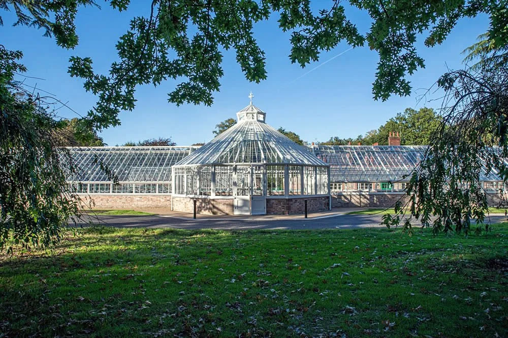 A large greenhouse with a glass roof, surrounded by trees and grass, under a clear blue sky.