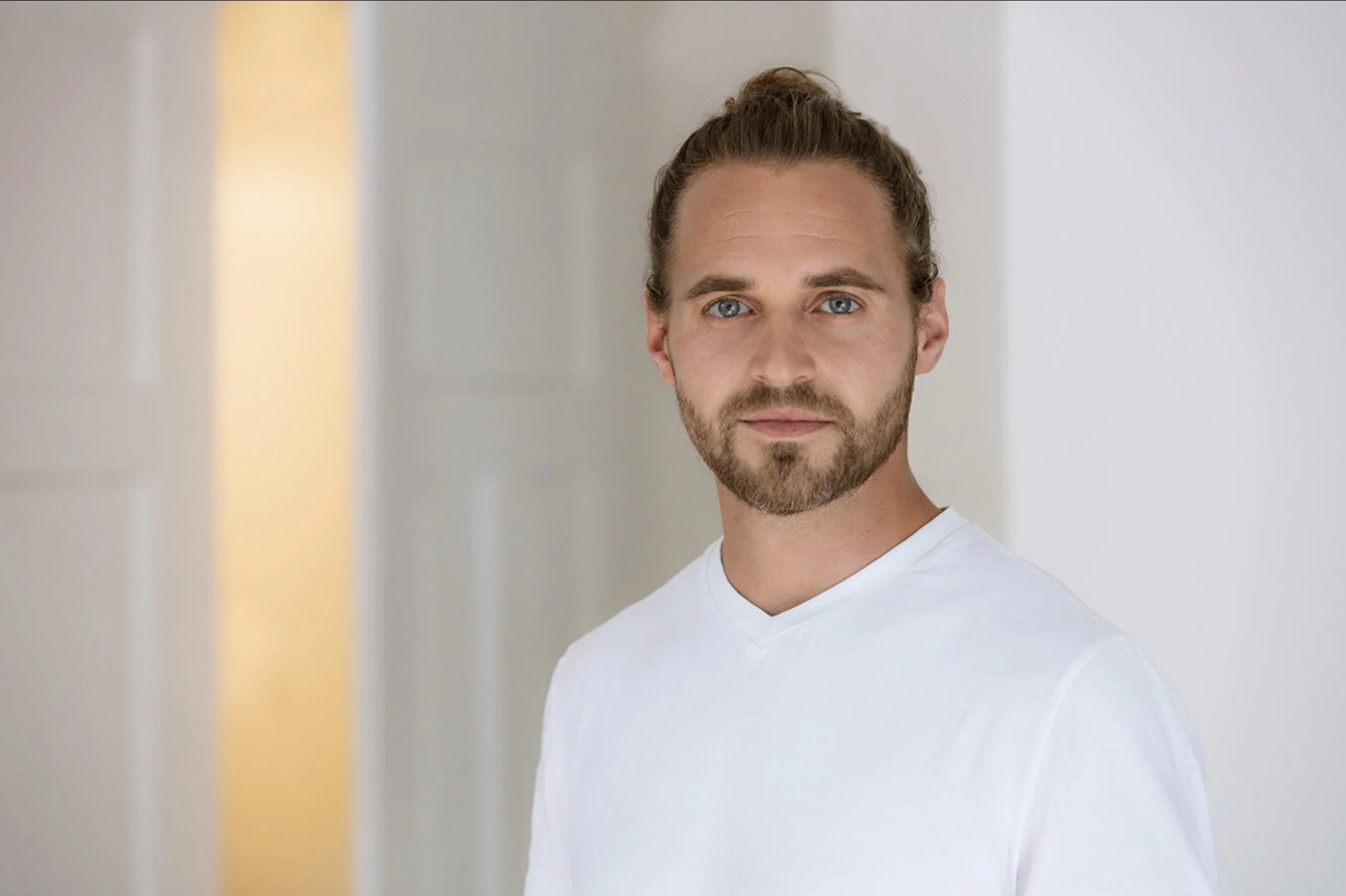 Junger Mann mit braunem Haar in einem weißen T-Shirt schaut in die Kamera, im Hintergrund eine helle, minimalistische Raumgestaltung.